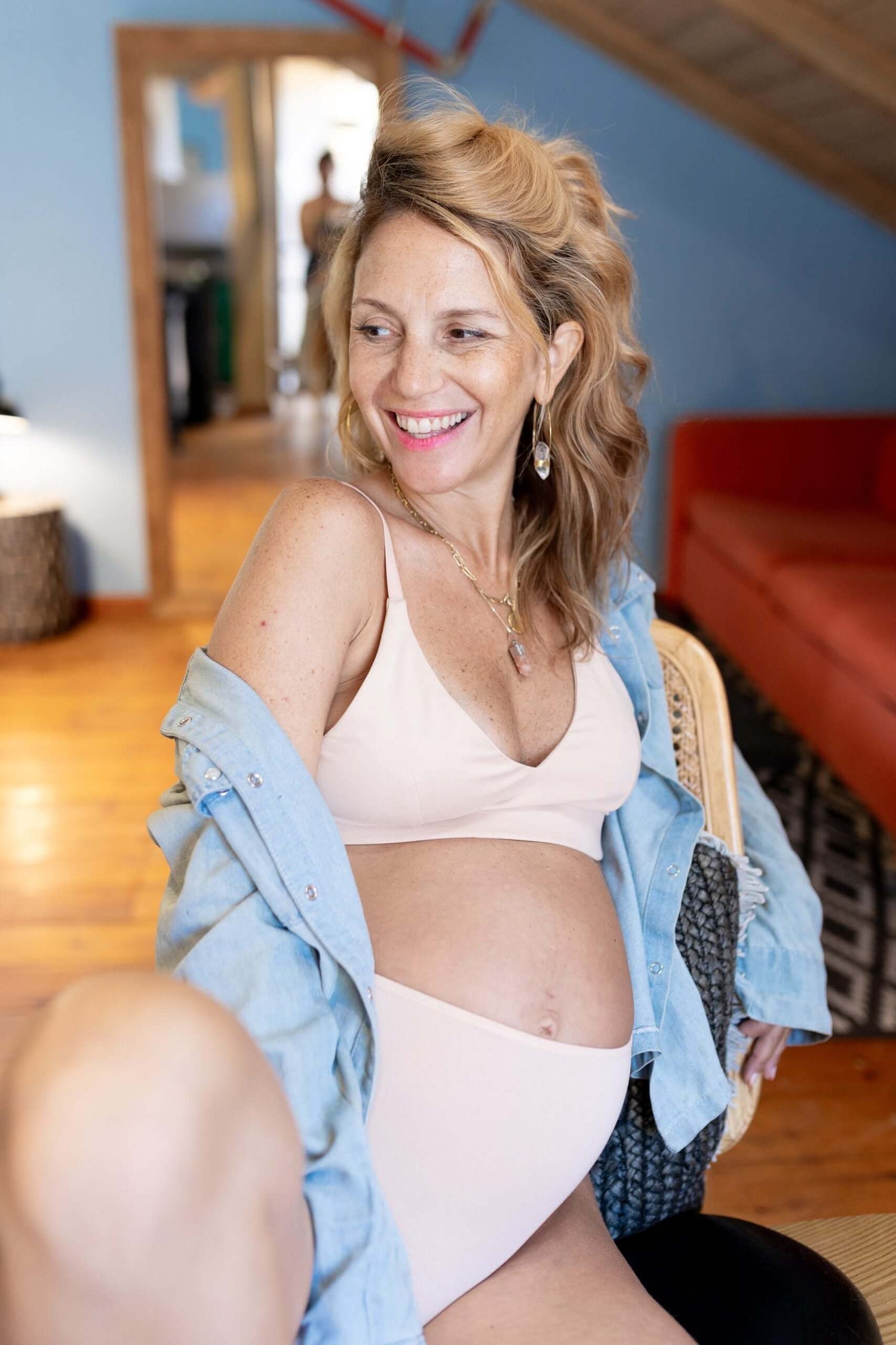 אישה בהריון שמחה בבית, חובשת חלוק כחול וחזיית ספורט, יושבת בנוחות בסלון.